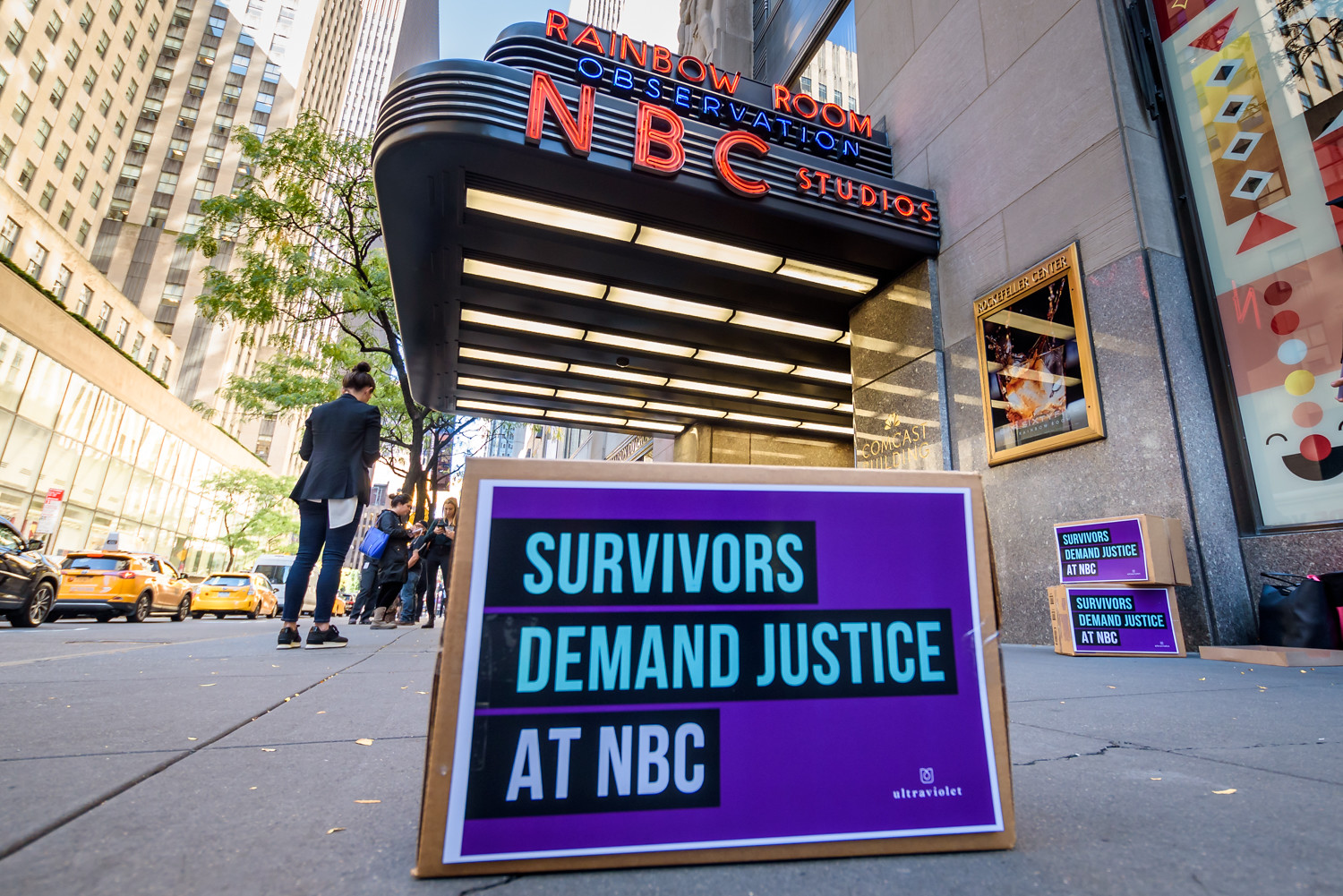 Box labeled "Survivors Demand Justice at NBC" sits in front of the exterior of NBC's 30 Rock