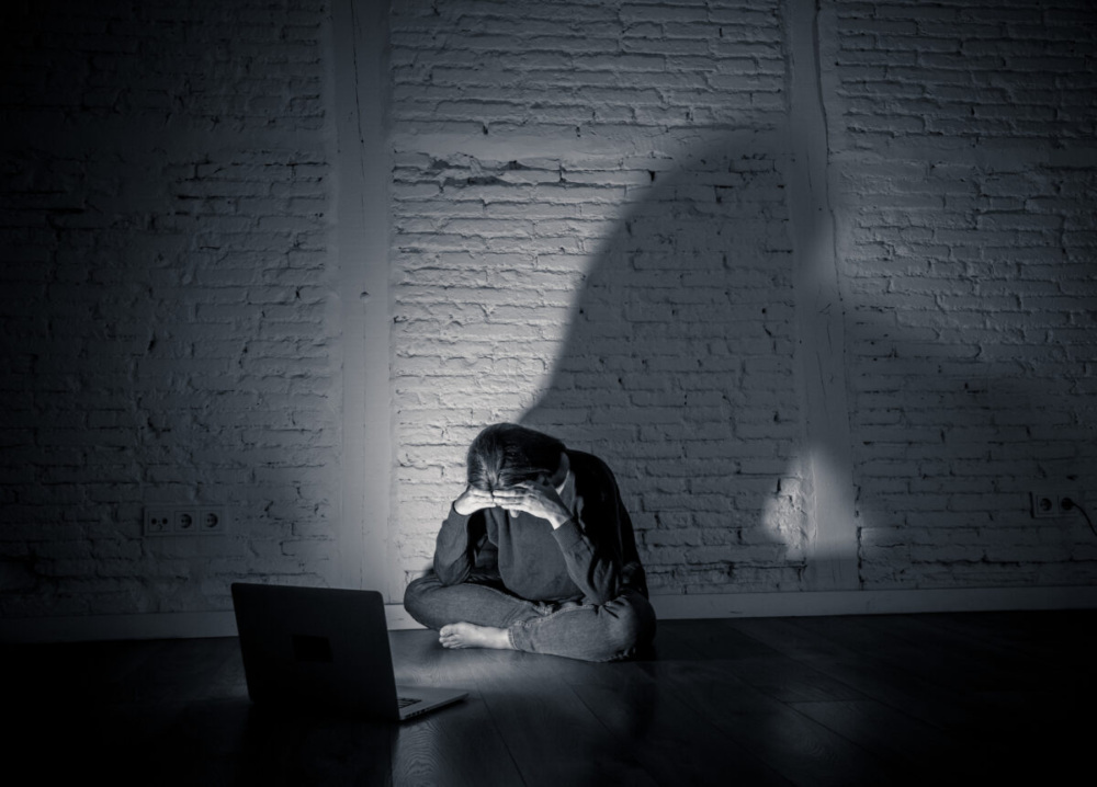 In a dimly lit room, a person sits on the floor facing an open laptop, their face buried in their hands.